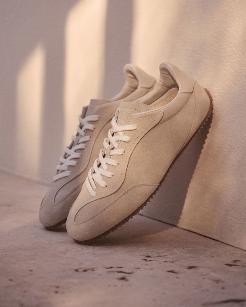 Beige Veloursleder-Sneaker mit weißen Schnürsenkeln und brauner Gummisohle, stehend an einer hellen Wand im Sonnenlicht.