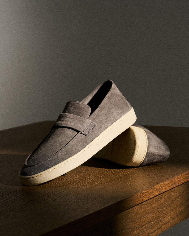Gray suede slip-on loafers with white rubber soles displayed on a wooden surface against a dark background.