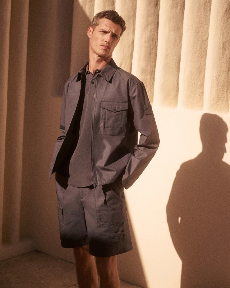 Man wearing a dark gray jacket and matching cargo shorts standing against a textured beige wall with hands in pockets.
