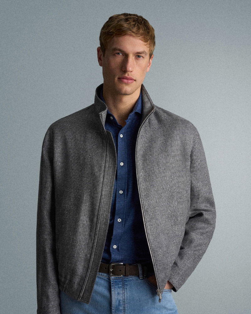 A man with short blond hair wears a gray zip-up jacket over a blue button-up shirt and light blue jeans, standing against a plain gray background.