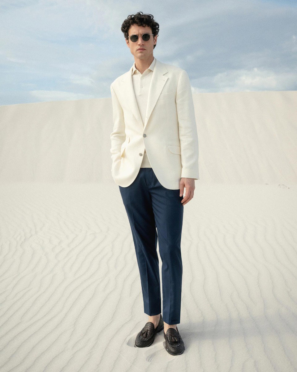 Man wearing round sunglasses, white blazer, cream shirt, navy trousers, and black loafers standing on white sand dunes.