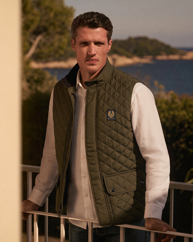 Man wearing green quilted vest and white shirt leaning on balcony railing with sea and trees in the background during sunset.