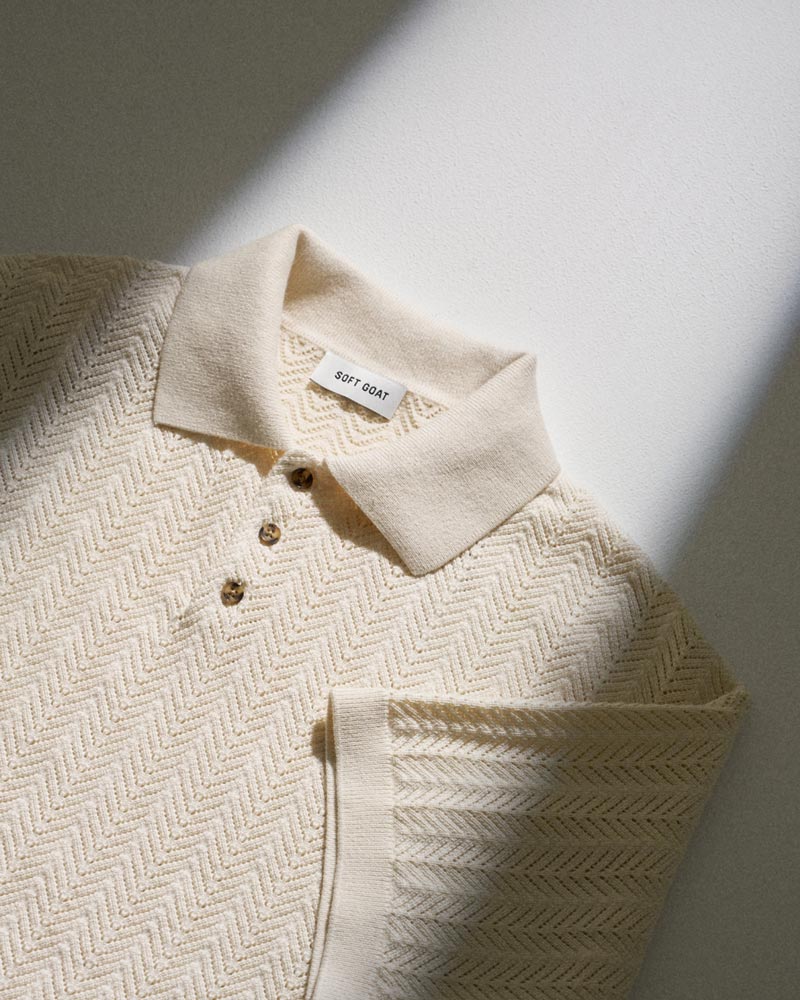 Cream-colored knit polo shirt with zigzag pattern, three buttons, and 'SOFT GOAT' label on collar, laid flat in sunlight.