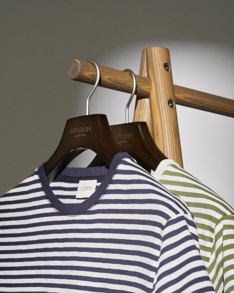 Two striped t-shirts, one navy and white and one green and white, hanging on dark wooden hangers on a wooden rack.