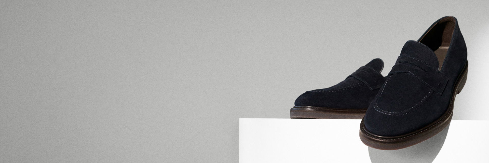 Dunkelblaue Wildleder-Herrenschuhe auf weißem Podest vor grauem Hintergrund, mit brauner Sohle und feiner Nahtverzierung. Dunkelblaue Wildleder-Herrenschuhe auf weißem Podest vor grauem Hintergrund, mit brauner Sohle und feiner Nahtverzierung.