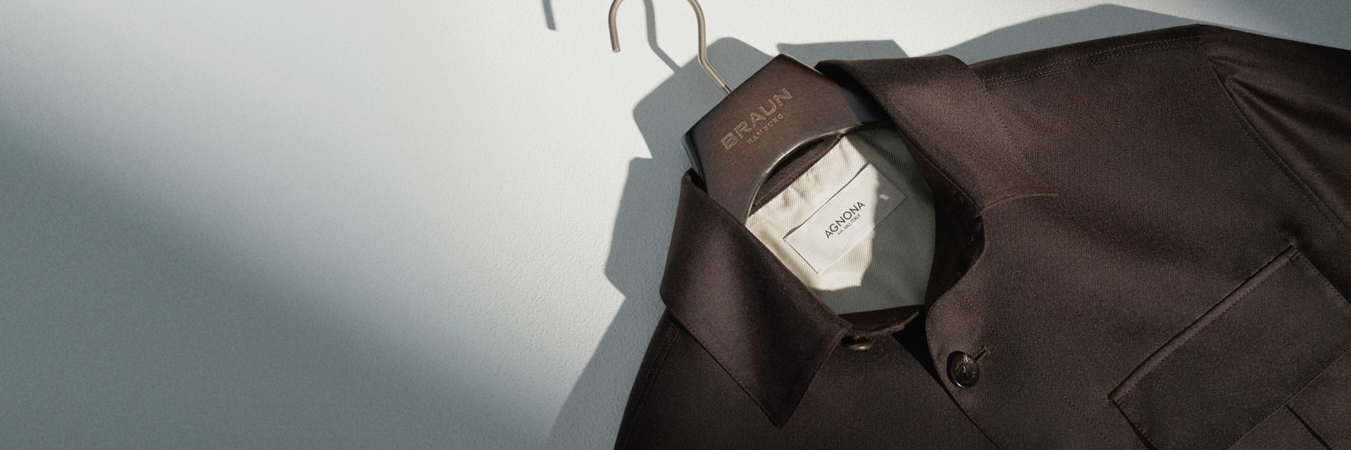 Dunkelbraune Jacke von Agnona auf einem Holzkleiderbügel mit Braun Hamburg Schriftzug vor weißer Wand. Dunkelbraune Jacke von Agnona auf einem Holzkleiderbügel mit Braun Hamburg Schriftzug vor weißer Wand.