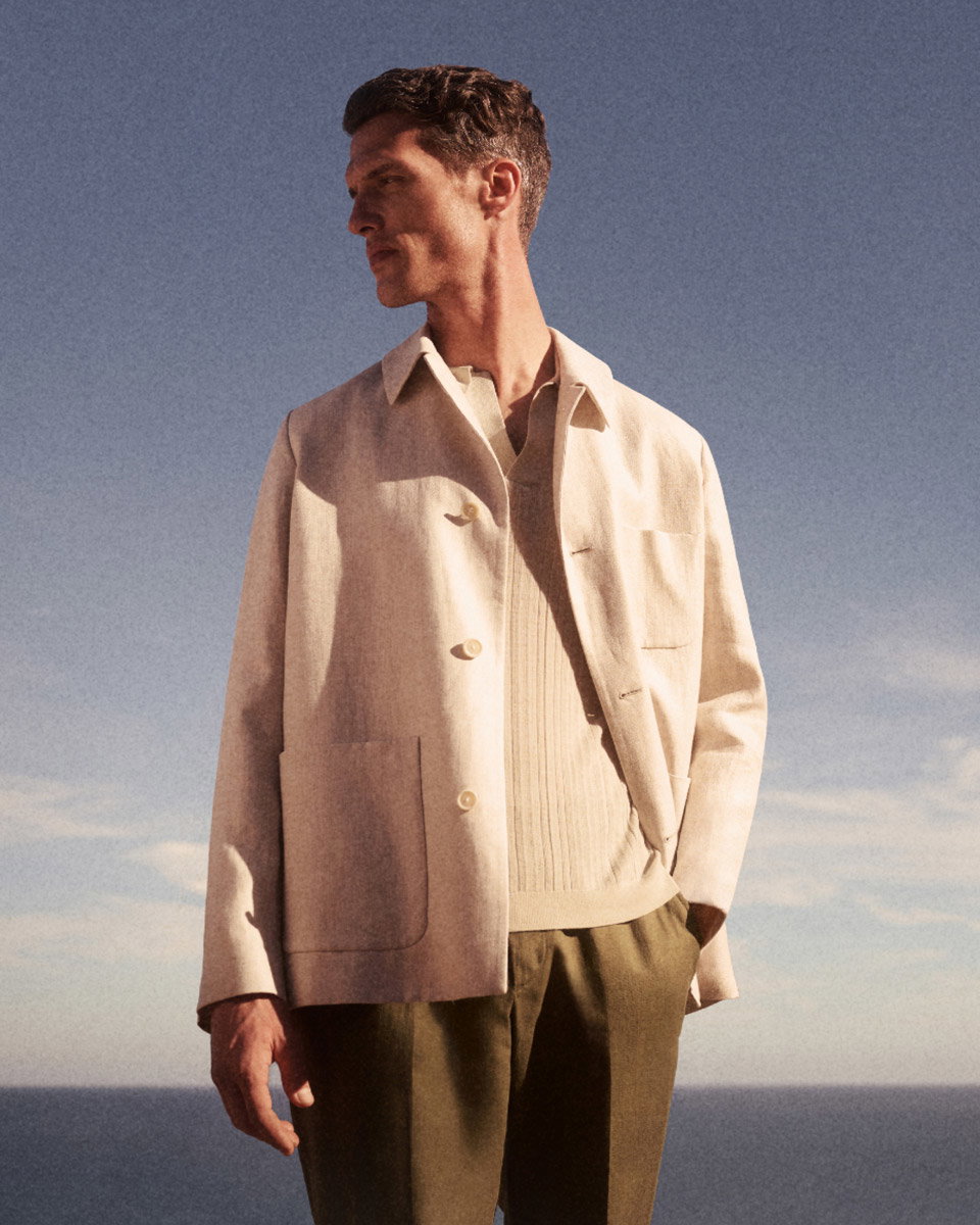 Mann mit kurzen braunen Haaren trägt beigen Mantel, hellen Pullover und olivgrüne Hose, steht vor blauem Himmel und Meer.