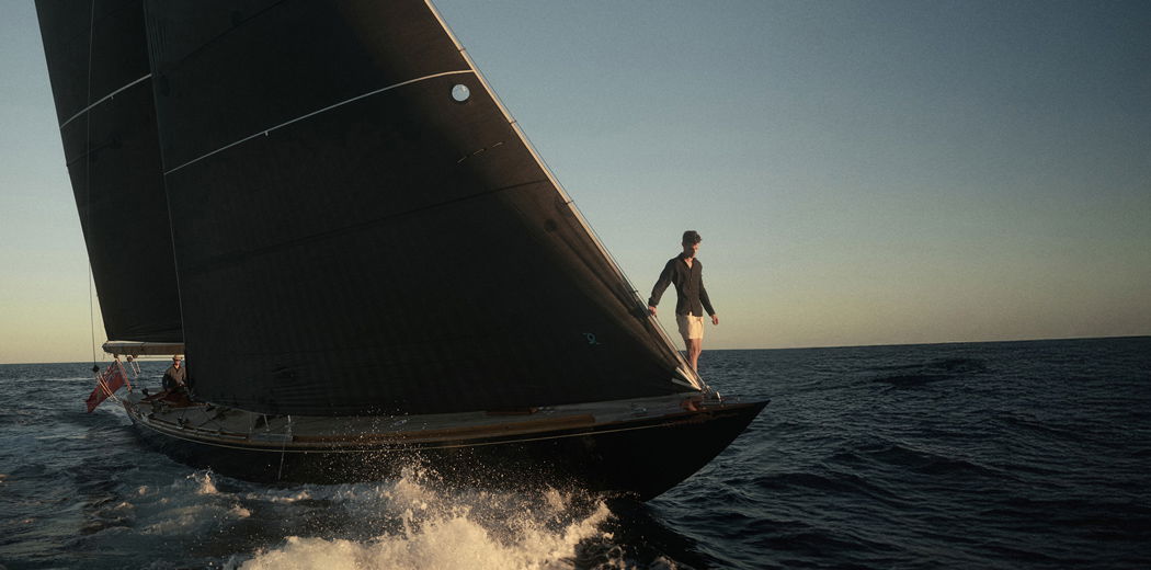 Eine Person steht vorne auf dem Bug eines schwarzen Segelboots, das bei Sonnenuntergang auf dem Meer segelt.