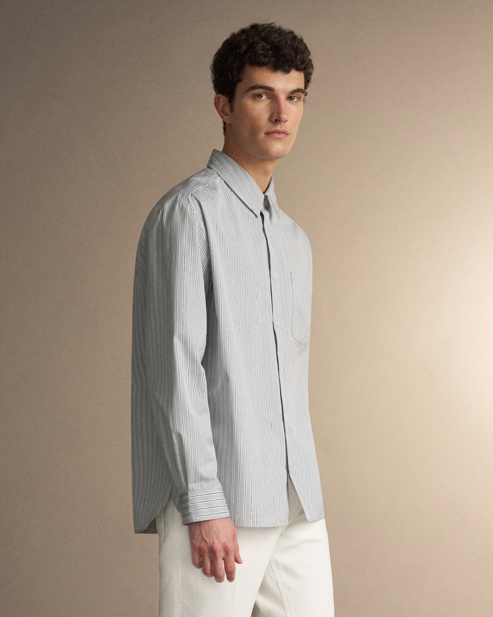 Junger Mann mit lockigem Haar trägt hellblaues, gestreiftes Casualhemd und weiße Hose vor beige Hintergrund.