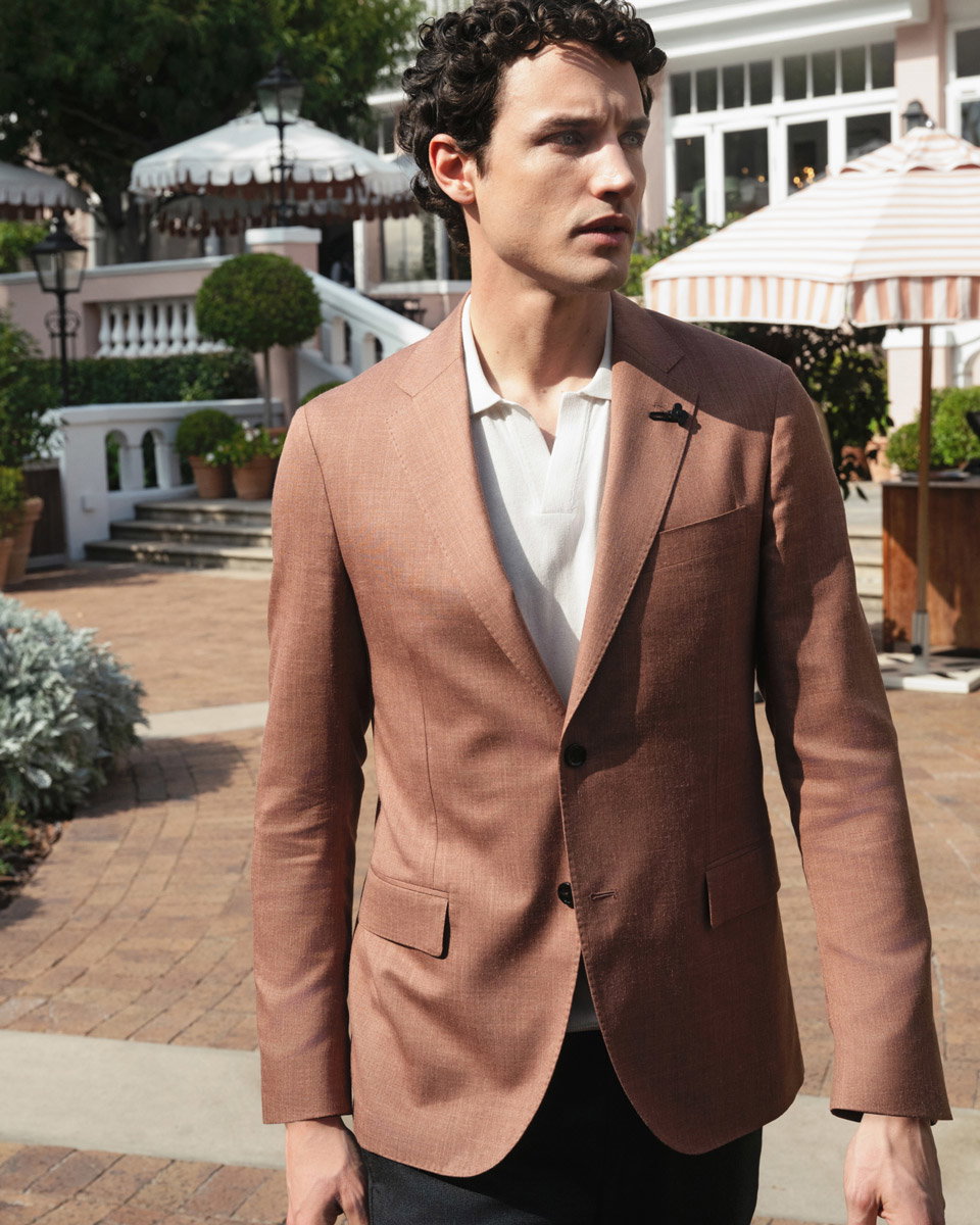 Mann mit braunem Sakko und weißem Poloshirt steht vor Terrasse mit Sonnenschirmen und Pflanzen, schaut nach rechts.