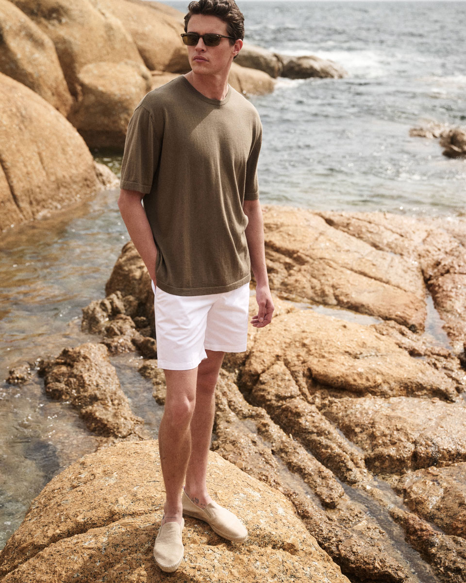 Mann mit Sonnenbrille, olivgrünem T-Shirt, weißen Shorts und Espadrilles steht auf Felsen am Meer bei sonnigem Wetter.