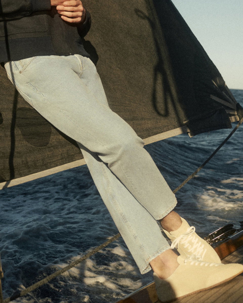 Person in hellblauer Jeans, cremefarbenen Sneakers und schwarzer Jacke lehnt auf Segelboot vor blauem Meer. Person in hellblauer Jeans, cremefarbenen Sneakers und schwarzer Jacke lehnt auf Segelboot vor blauem Meer.