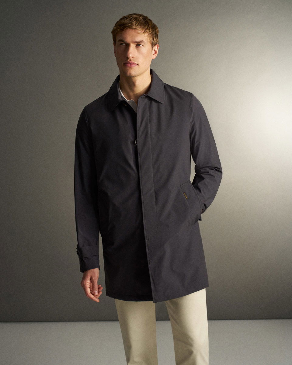 Mann mit blondem Haar trägt einen dunkelblauen Mantel und helle Hose vor schlichtem grauem Hintergrund.