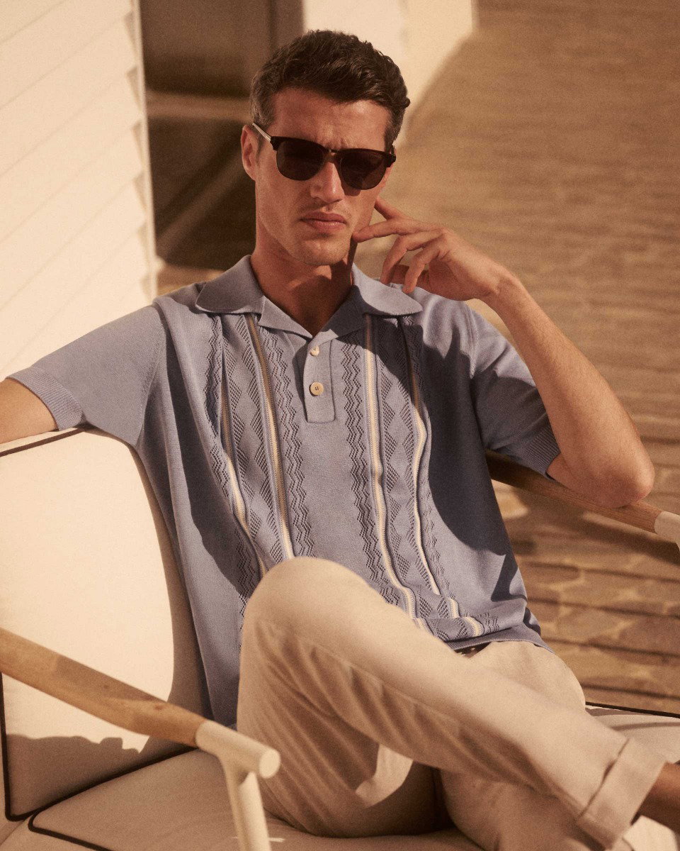 Mann mit dunkelbraunen Haaren trägt blaue gemusterte Polohemd, helle Hose und schwarze Sonnenbrille, sitzt entspannt draußen.