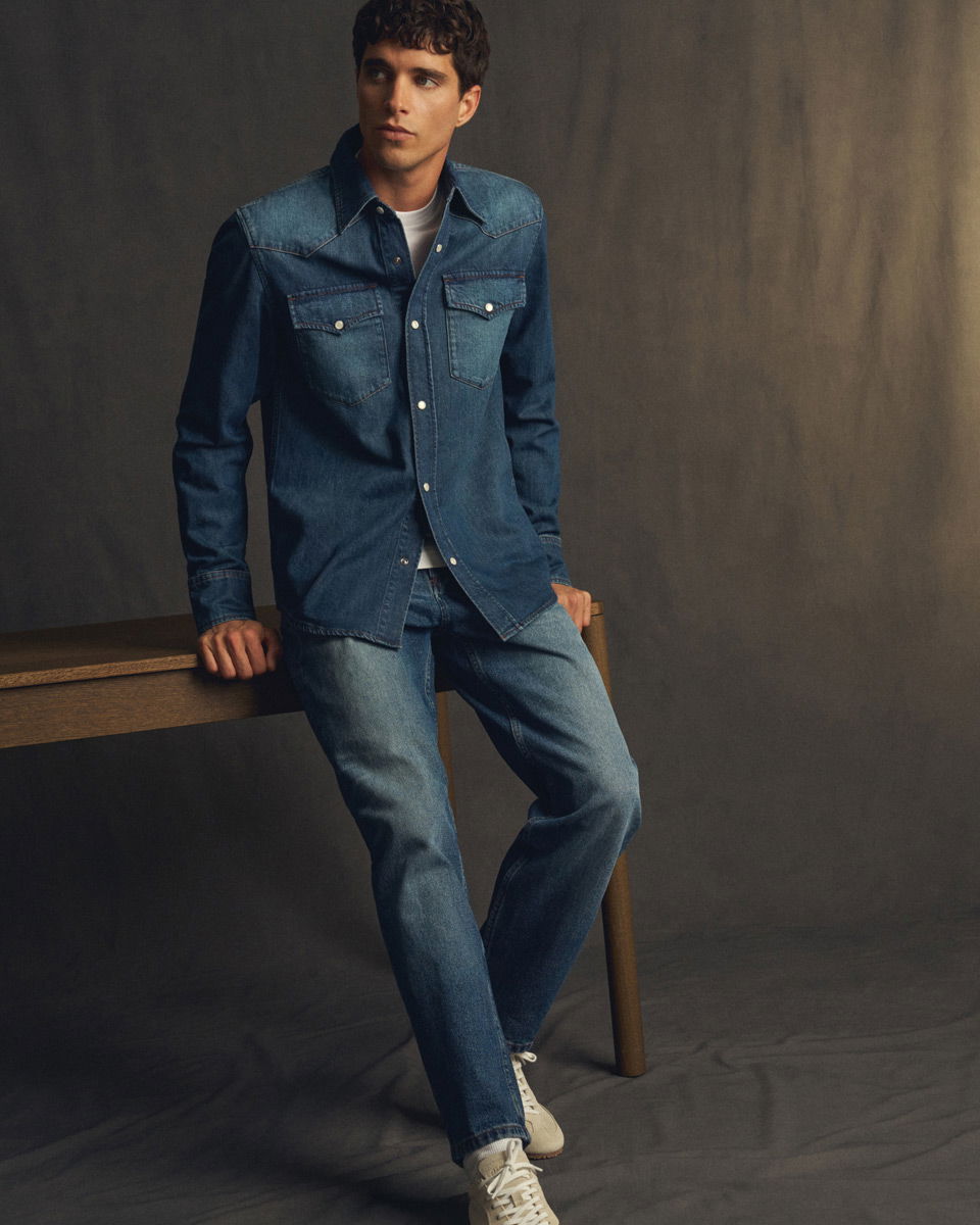 Mann lehnt an Holztisch, trägt dunkelblaue Jeansjacke, blaue Jeans und weiße Sneaker vor grauem Hintergrund.