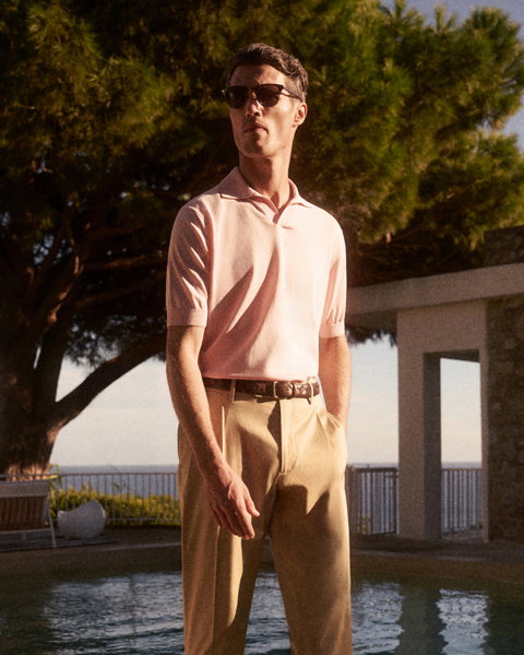 Mann mit Sonnenbrille trägt hellrosa Poloshirt und beige Hose, steht vor Pool mit Bäumen und Meer im Hintergrund.