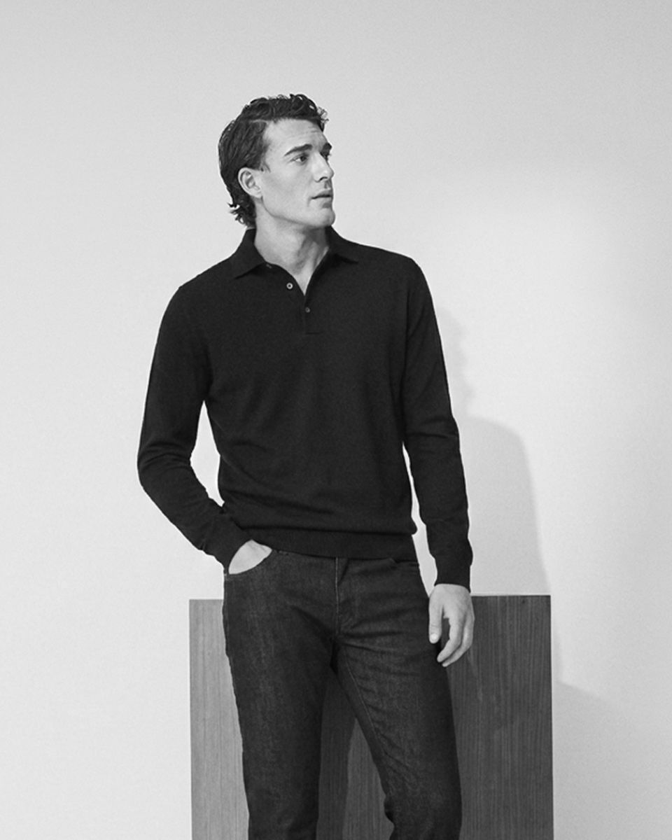 Mann in schwarzem Pullover und Jeans steht vor weißer Wand, linke Hand in Tasche, schaut nach rechts, Schwarz-Weiß-Foto.