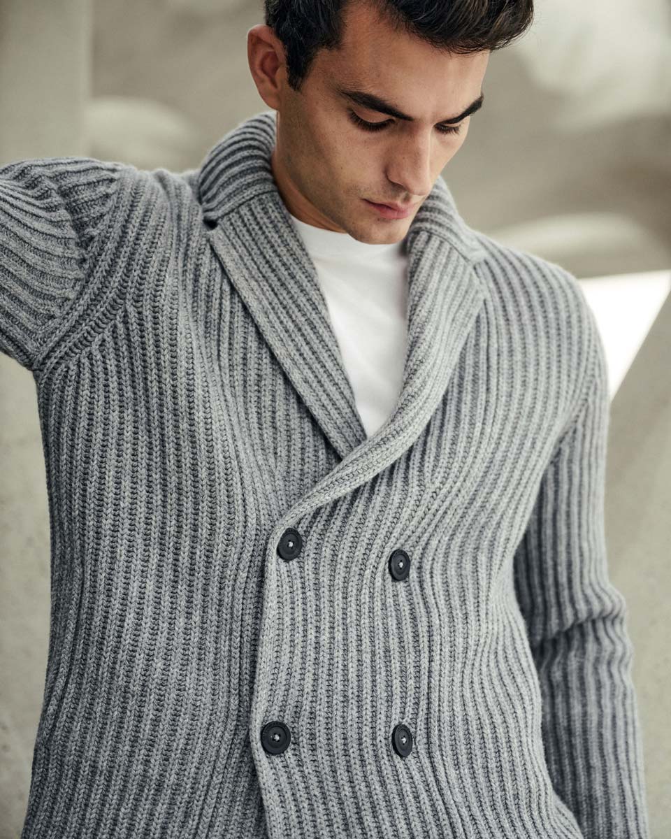 Mann trägt hellgrauen, grob gestrickten, doppelt geknöpften Cardigan mit breitem Kragen über weißem T-Shirt, nach unten schauend. Mann trägt hellgrauen, grob gestrickten, doppelt geknöpften Cardigan mit breitem Kragen über weißem T-Shirt, nach unten schauend.