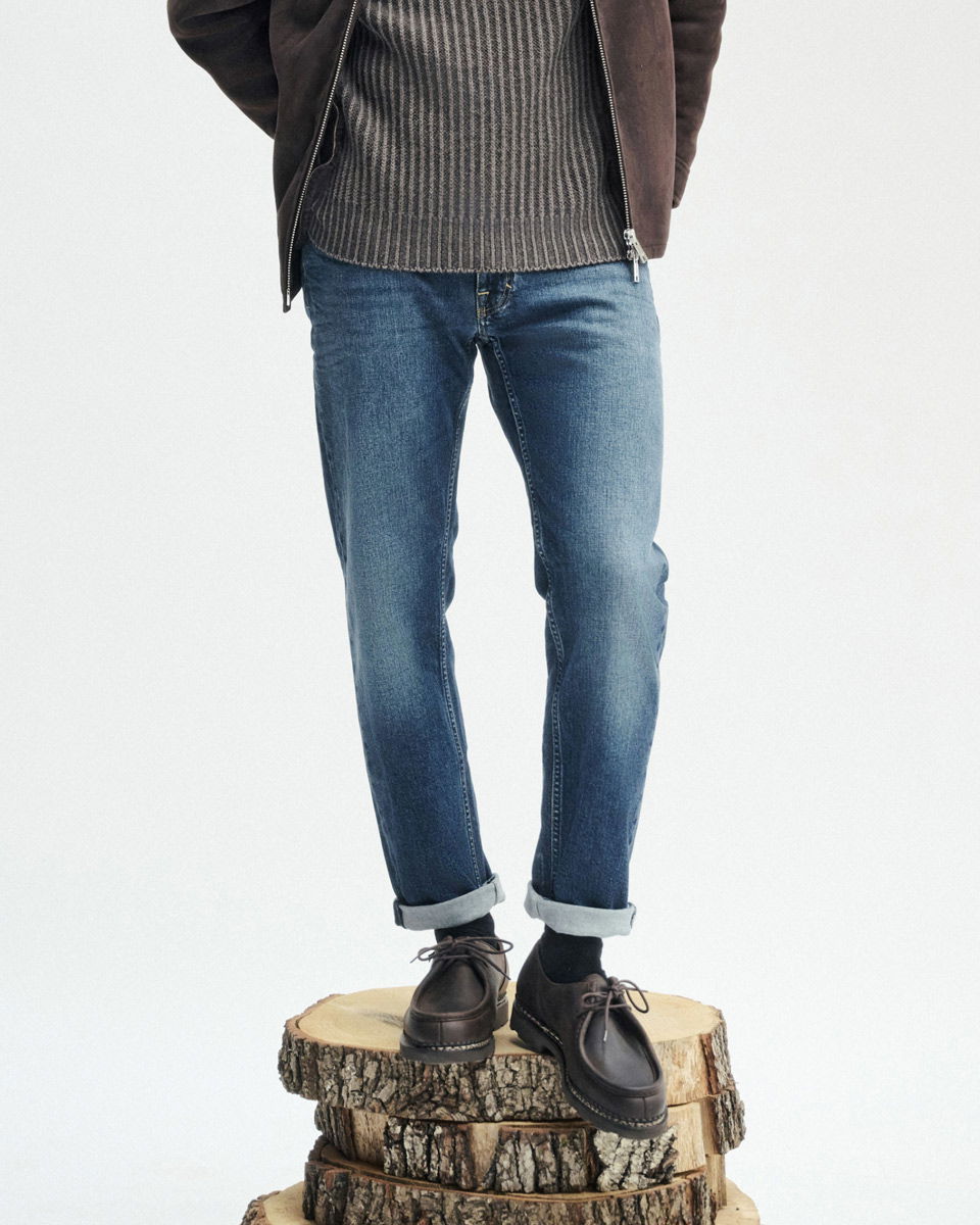 Mann trägt dunkelbraune Schuhe, dunkelblaue Jeans mit Umschlag, grauen Pullover und braune Jacke, steht auf Baumstamm.