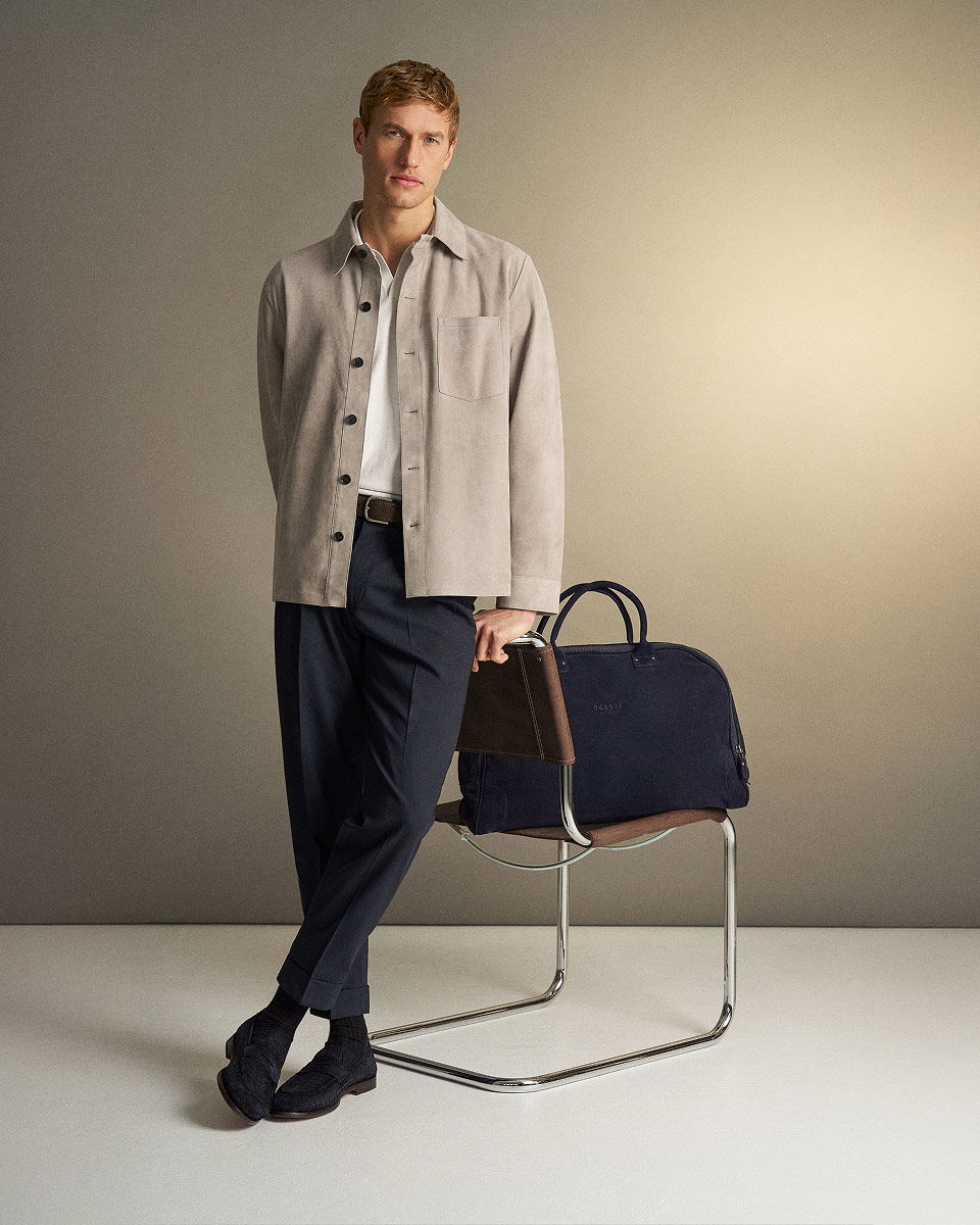 Junger Mann lehnt an Stuhl mit dunkelblauer Reisetasche, trägt graue Jacke, weiße Bluse, dunkelblaue Hose und schwarze Schuhe.