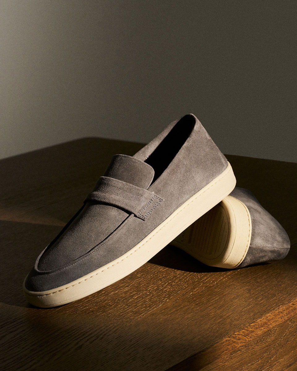 Grauer Veloursleder-Slipper mit cremefarbener Gummisohle, auf Holztisch stehend, in warmem Licht und Schatten.