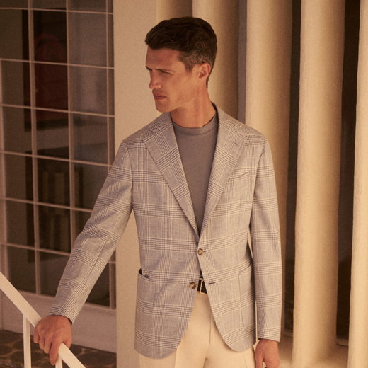 Mann mit braunen Haaren trägt kariertes Sakko, graues Shirt und helle Hose, steht vor Säulen und schaut zur Seite.