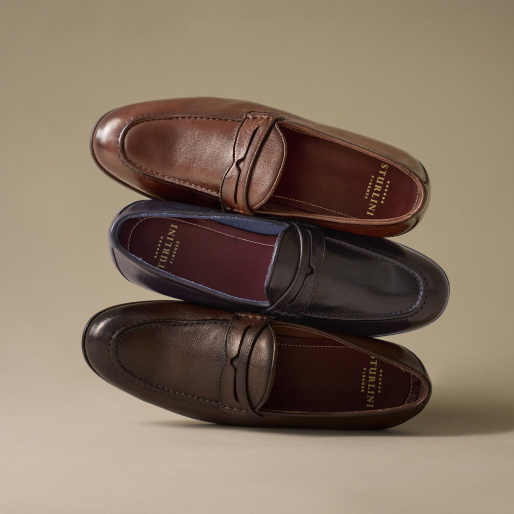 Drei hochwertige Stürlini Lederschuhe in Braun, Dunkelbraun und Blau, elegant gestapelt vor beigem Hintergrund.