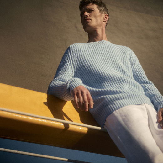 Mann in hellblauem Strickpullover und weißer Hose lehnt entspannt an gelbem Geländer vor brauner Wand im Sonnenlicht.