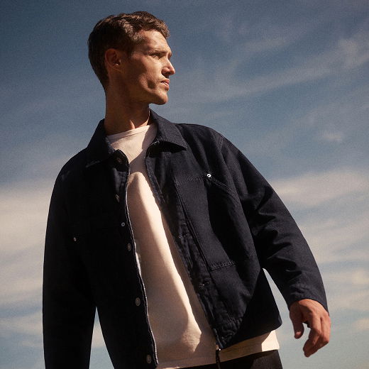 Junger Mann mit kurzem Haar trägt weiße Shirt und schwarze Jacke, blickt nach links, blauer Himmel mit Wolken im Hintergrund.