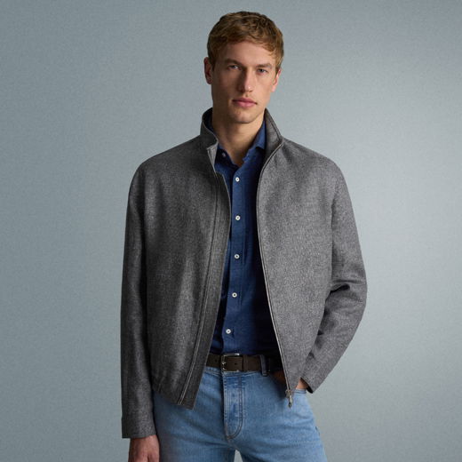 Junger Mann mit blonden Haaren trägt graue Jacke über blauem Hemd und hellblauen Jeans, steht vor grauem Hintergrund.