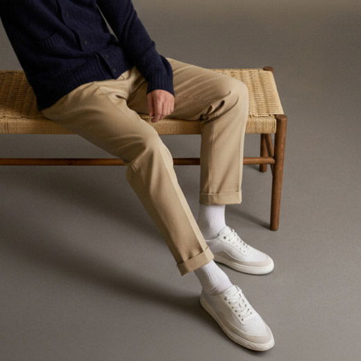 Mann sitzt auf geflochtenem Holzbank, trägt beige Hose, weiße Sneakers, weiße Socken und dunkelblauen Pullover, grauer Hintergrund.