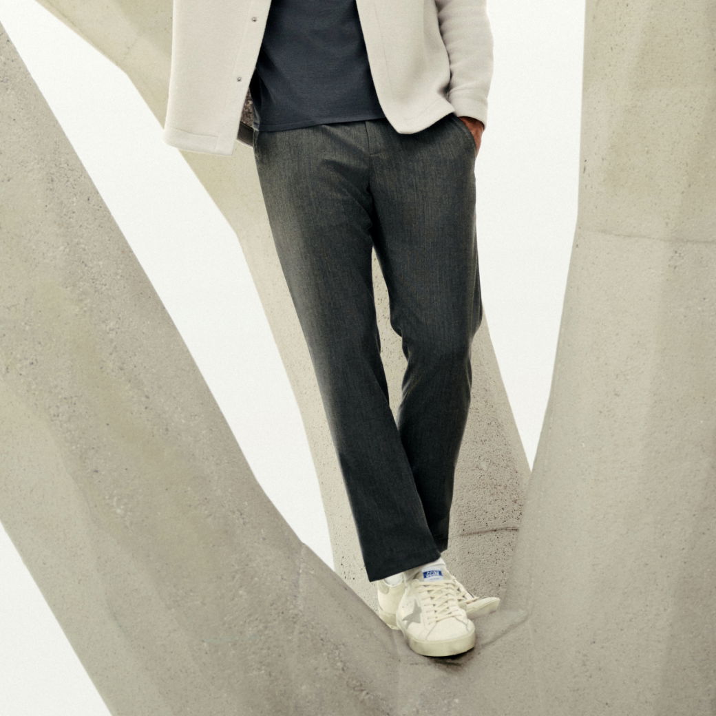 Mann trägt dunkelgraue Wollhose, weiße Sneaker und hellen Mantel, steht zwischen großen Betonstrukturen. Mann trägt dunkelgraue Wollhose, weiße Sneaker und hellen Mantel, steht zwischen großen Betonstrukturen.