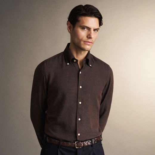 Junger Mann mit dunklen Haaren trägt braunes Hemd und Ledergürtel, steht vor beige Hintergrund, schaut ernst in Kamera.