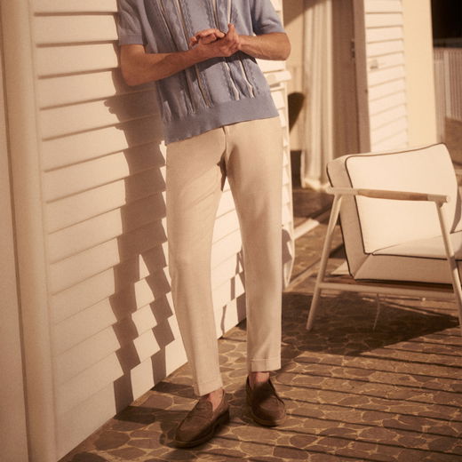 Mann lehnt an weißer Hauswand, trägt beige Hose, hellblauen Pullover und braune Loafers, neben ihm moderner weißer Stuhl.