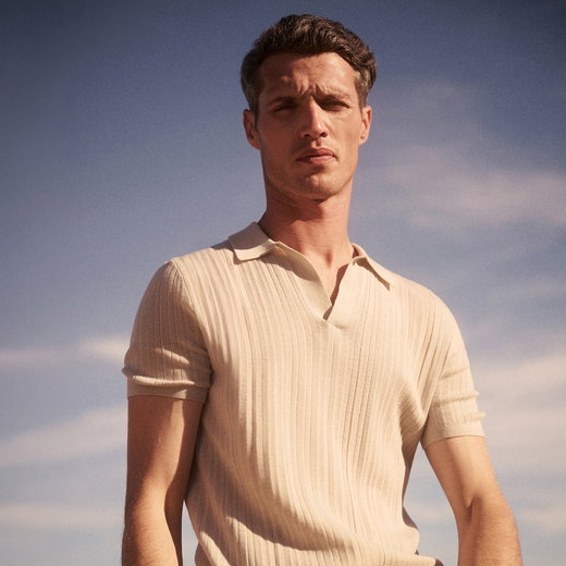 Junger Mann mit kurzen braunen Haaren trägt hellbeiges, kurzärmeliges, geripptes Poloshirt vor blauem Himmel.