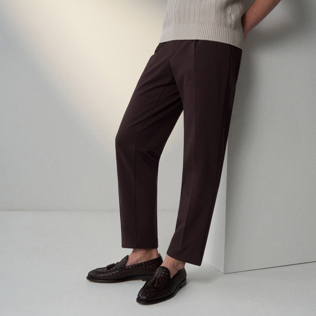 Dunkelbraune, elegante Hose mit Bügelfalte, kombiniert mit dunkelbraunen Tassel-Loafern, sitzend an einer hellen Wand.