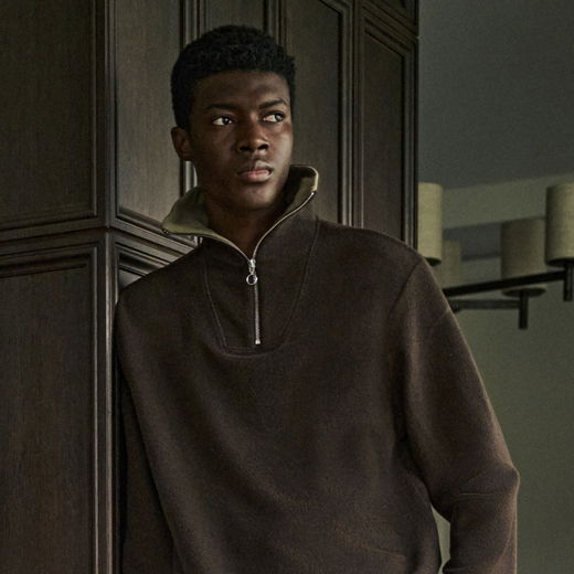 Junger Mann mit kurzem Afrohaarschnitt trägt braunen Troyer-Pullover mit Reißverschluss, lehnt an dunklem Holzschrank.