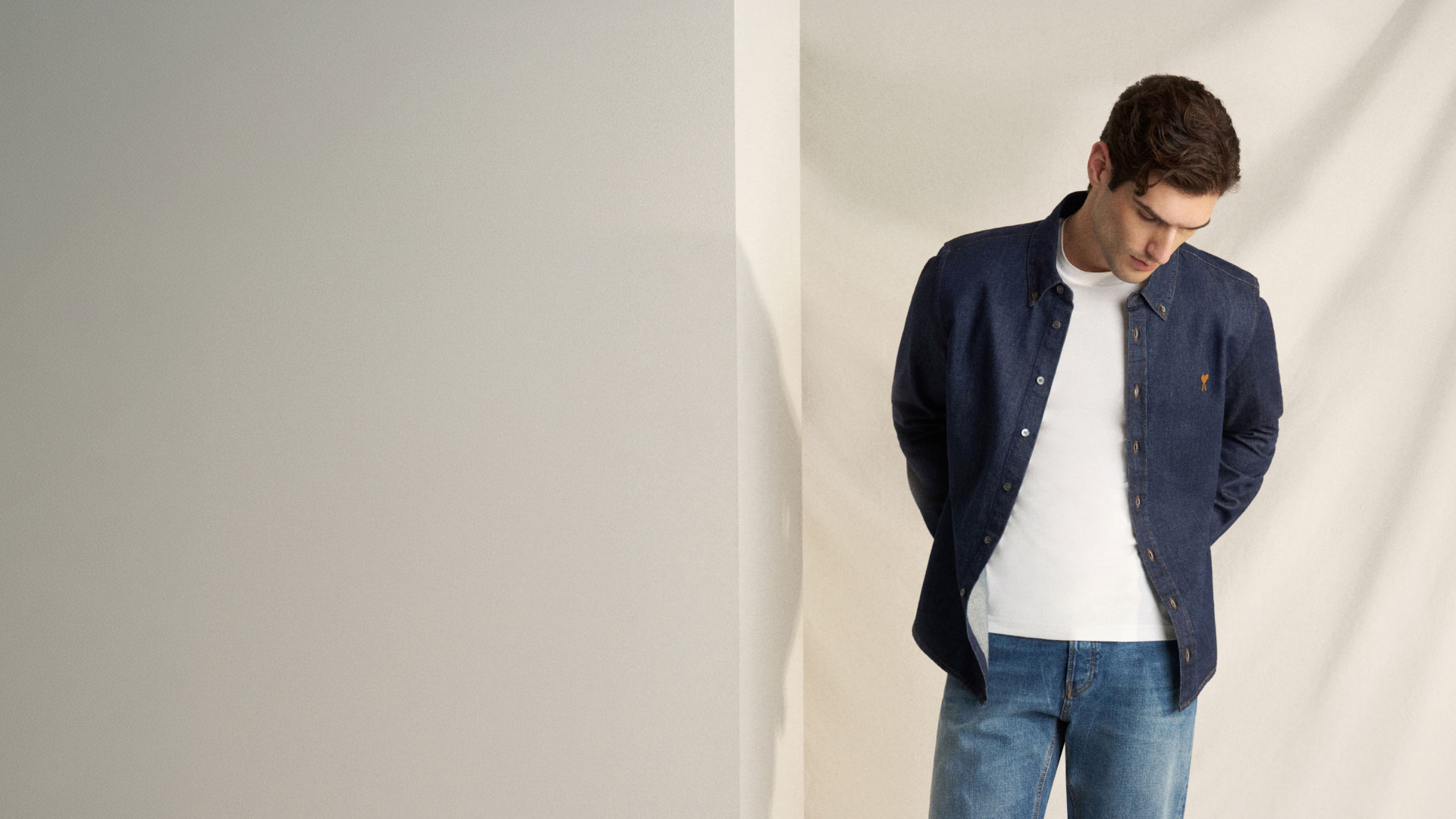 Mann trägt weiße T-Shirt, offene dunkelblaue Jeansjacke und blaue Jeans, schaut nach unten vor hellem Hintergrund. Mann trägt weiße T-Shirt, offene dunkelblaue Jeansjacke und blaue Jeans, schaut nach unten vor hellem Hintergrund.