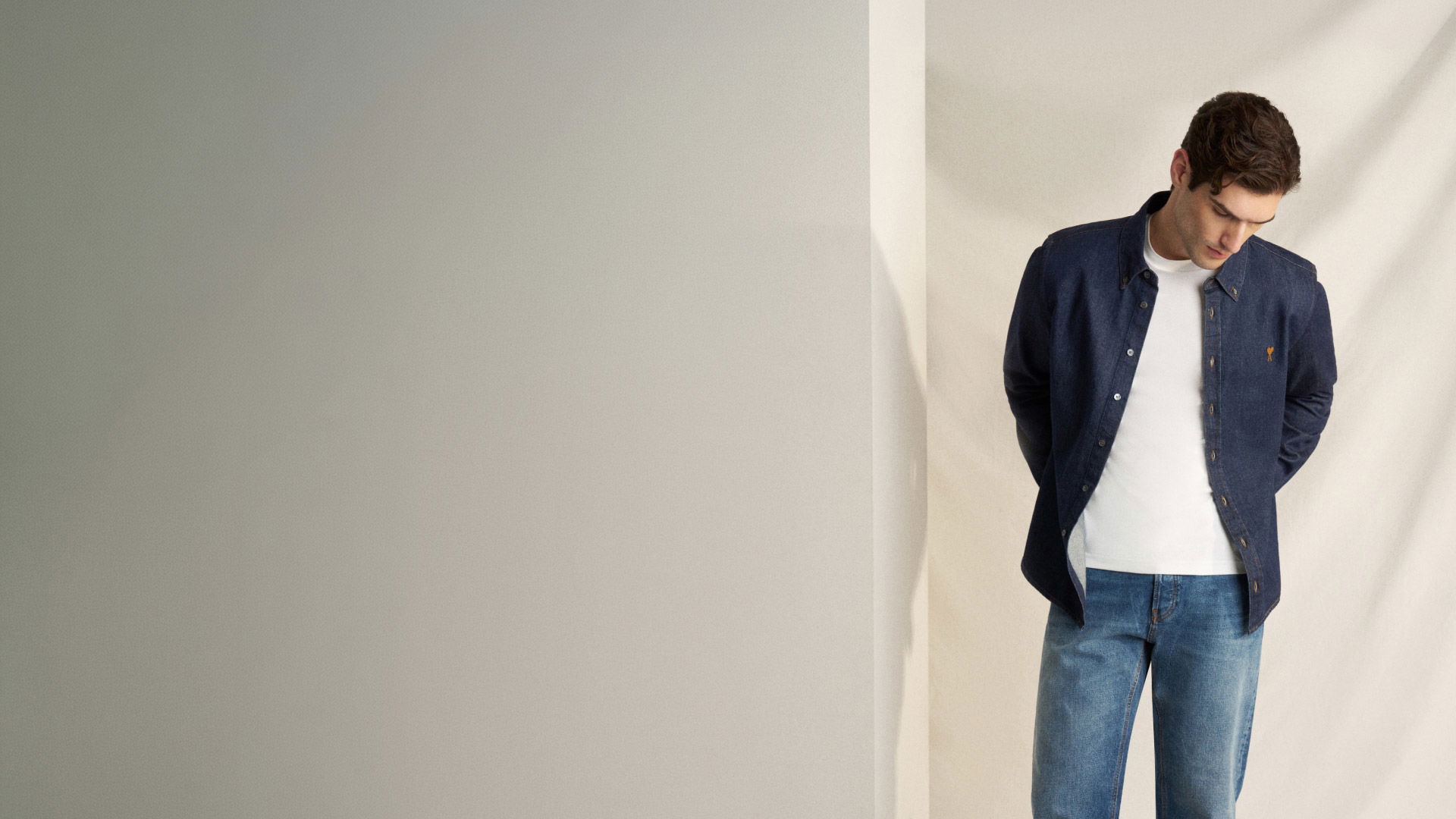 Young man wearing a white t-shirt, dark blue buttoned shirt, and blue jeans, standing against a beige wall looking down. Young man wearing a white t-shirt, dark blue buttoned shirt, and blue jeans, standing against a beige wall looking down.