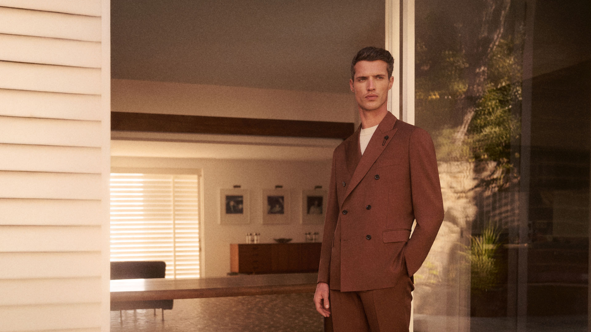 Mann in braunem Anzug steht vor modernem Haus mit großen Glasfenstern und Innenraum mit Möbeln und Bildern. Mann in braunem Anzug steht vor modernem Haus mit großen Glasfenstern und Innenraum mit Möbeln und Bildern.