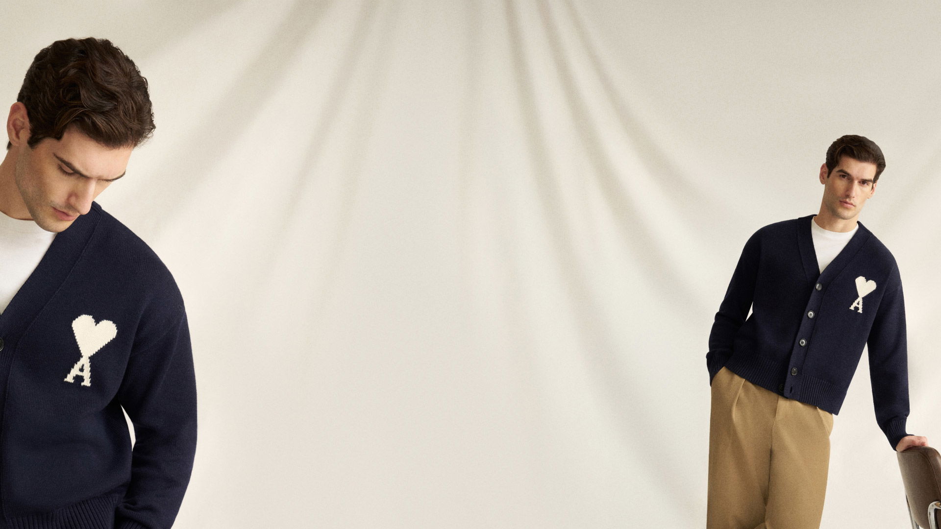 Junger Mann mit dunklem Cardigan mit weißem Herz-Logo und beiger Hose lehnt an Stuhl vor hellem Hintergrund. Junger Mann mit dunklem Cardigan mit weißem Herz-Logo und beiger Hose lehnt an Stuhl vor hellem Hintergrund.