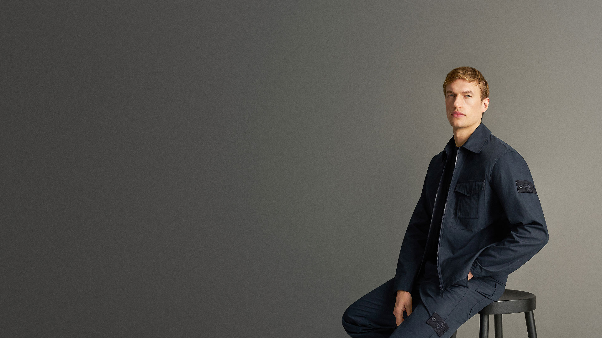 Junger Mann sitzt auf schwarzem Hocker, trägt dunkelblaue Jacke und Hose vor grauem Hintergrund, schaut direkt in Kamera. Junger Mann sitzt auf schwarzem Hocker, trägt dunkelblaue Jacke und Hose vor grauem Hintergrund, schaut direkt in Kamera.