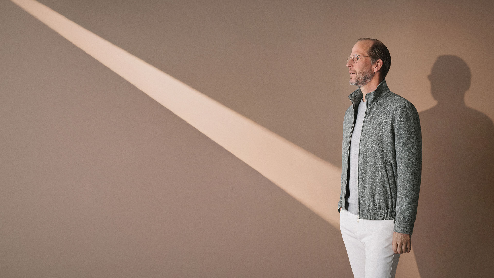 Mann mit Brille und grauer Jacke steht vor beiger Wand, Sonnenstrahl fällt schräg auf die Wand, Schatten sichtbar. Mann mit Brille und grauer Jacke steht vor beiger Wand, Sonnenstrahl fällt schräg auf die Wand, Schatten sichtbar.