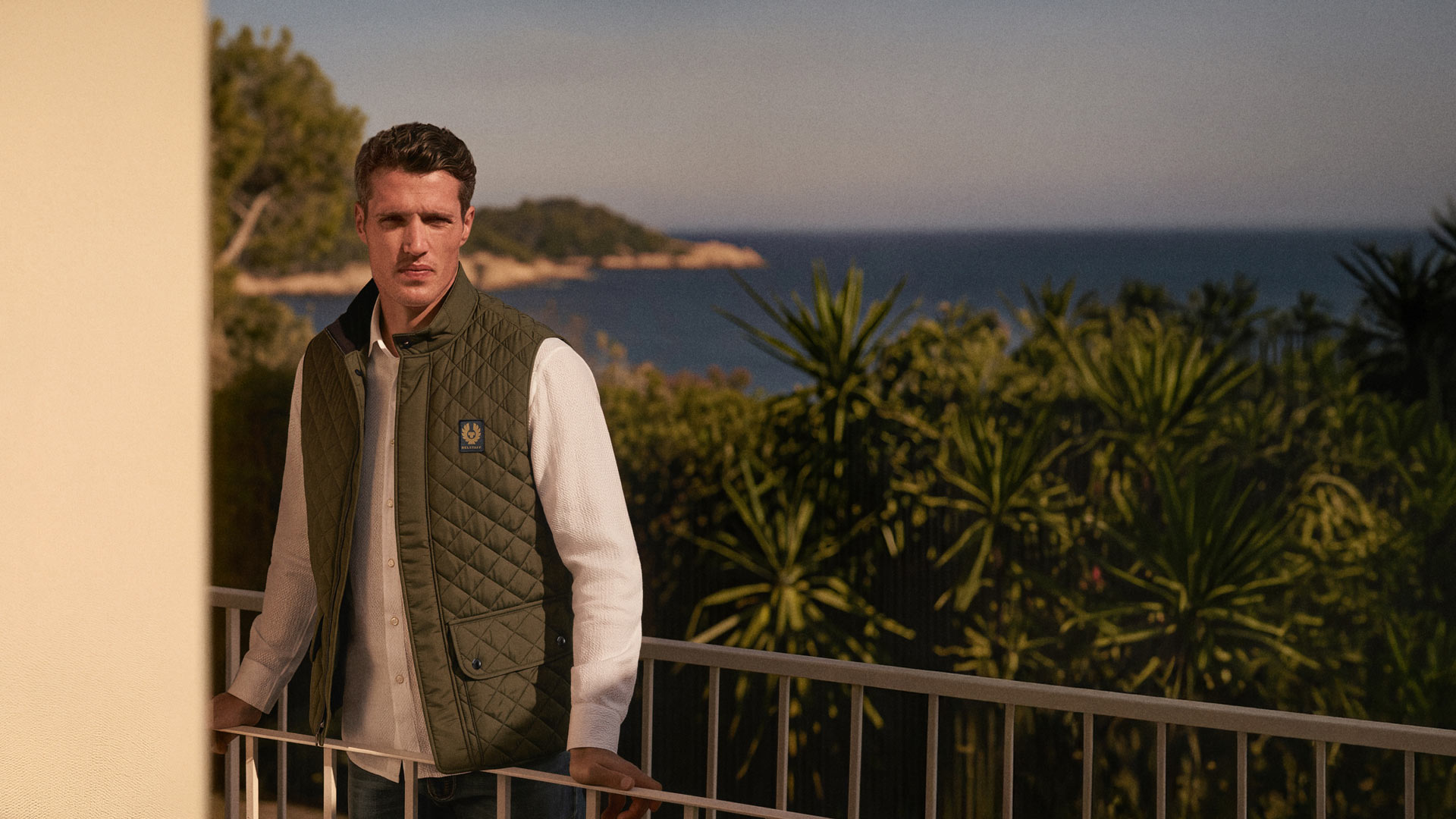 Mann mit weißem Hemd und grüner gesteppter Weste lehnt auf Balkon, im Hintergrund Meer, Bäume und blauer Himmel. Mann mit weißem Hemd und grüner gesteppter Weste lehnt auf Balkon, im Hintergrund Meer, Bäume und blauer Himmel.