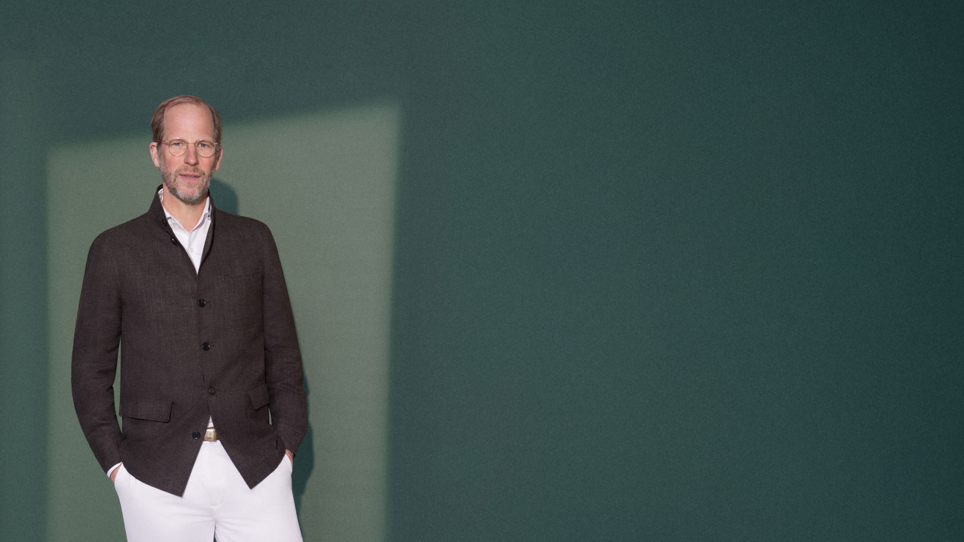 Mann mit Brille trägt braune Jacke und weiße Hose, steht vor grünem Hintergrund mit Sonnenlicht-Schatten. Mann mit Brille trägt braune Jacke und weiße Hose, steht vor grünem Hintergrund mit Sonnenlicht-Schatten.