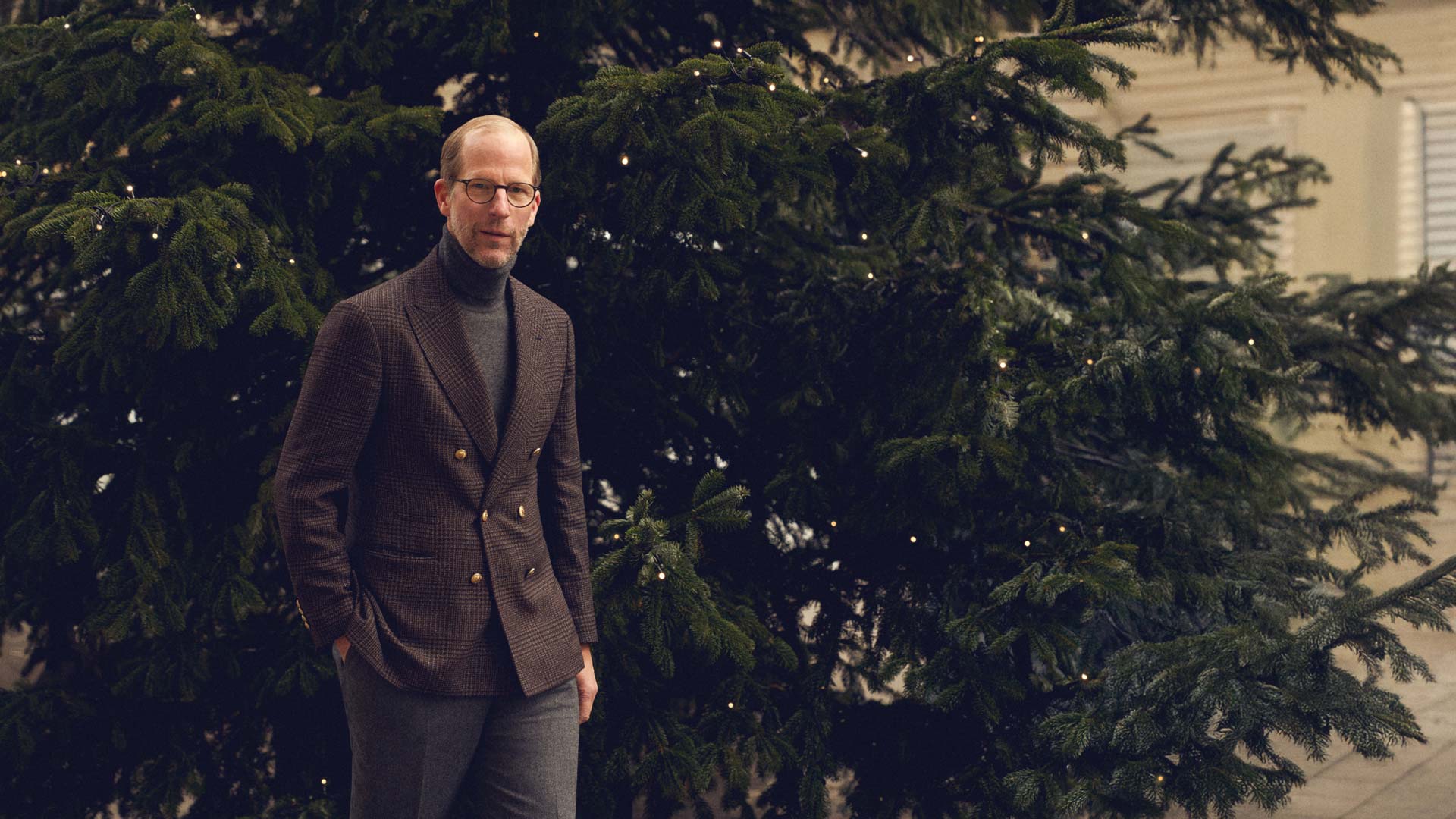 Mann mit Brille, braunem Sakko und grauem Rollkragenpullover steht vor großem Tannenbaum mit Lichterkette, Hände in Taschen. Mann mit Brille, braunem Sakko und grauem Rollkragenpullover steht vor großem Tannenbaum mit Lichterkette, Hände in Taschen.