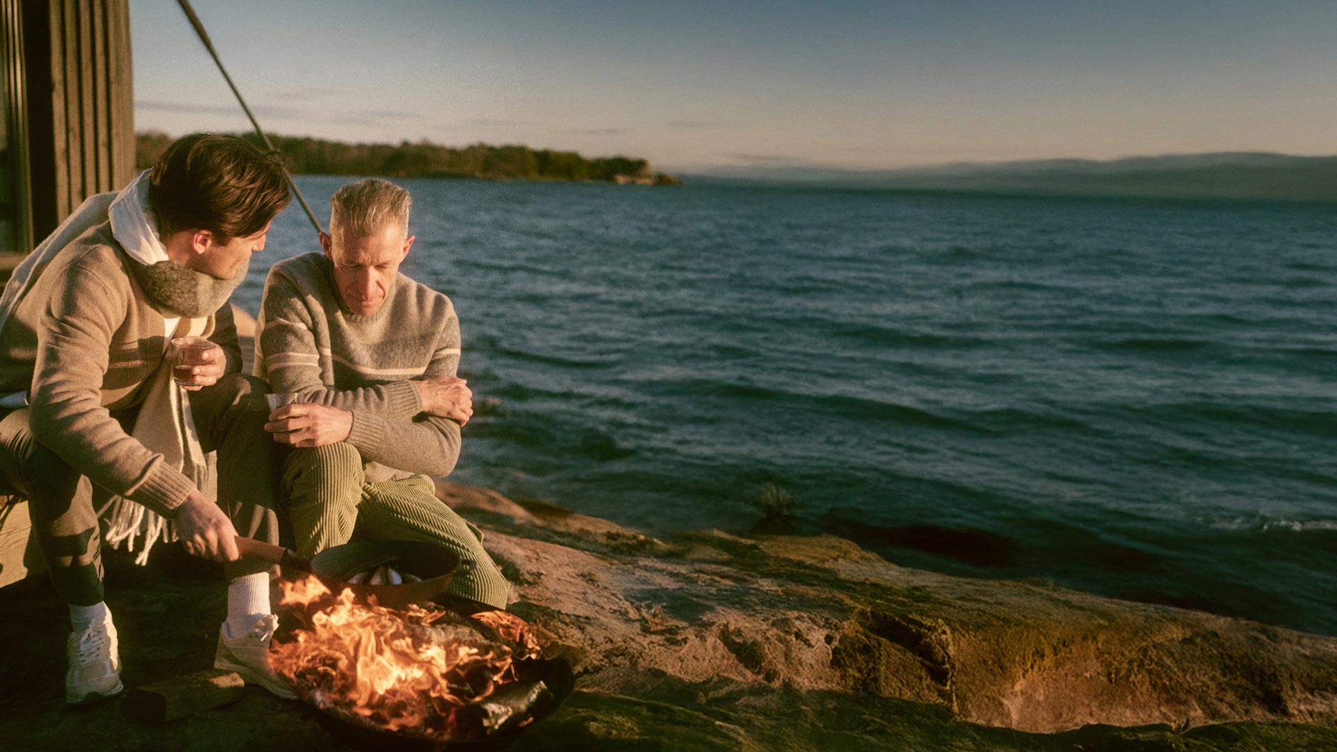 Zwei Männer sitzen am Wasser auf einem Felsen, wärmen sich an einem offenen Feuer und halten Getränke in der Hand. Zwei Männer sitzen am Wasser auf einem Felsen, wärmen sich an einem offenen Feuer und halten Getränke in der Hand.