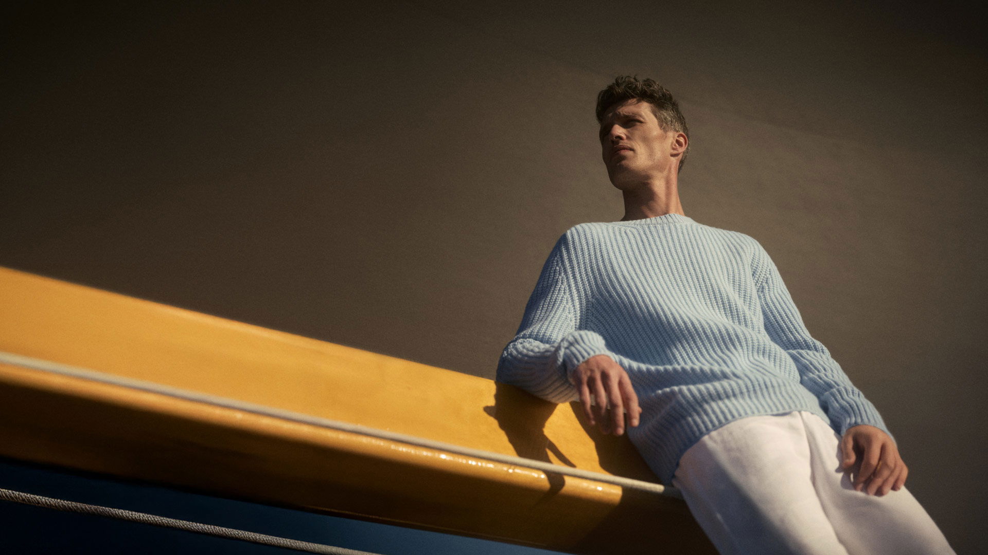 Mann in hellblauem Strickpullover und weißer Hose lehnt an gelbem Geländer vor dunkelbraunem Hintergrund. Mann in hellblauem Strickpullover und weißer Hose lehnt an gelbem Geländer vor dunkelbraunem Hintergrund.