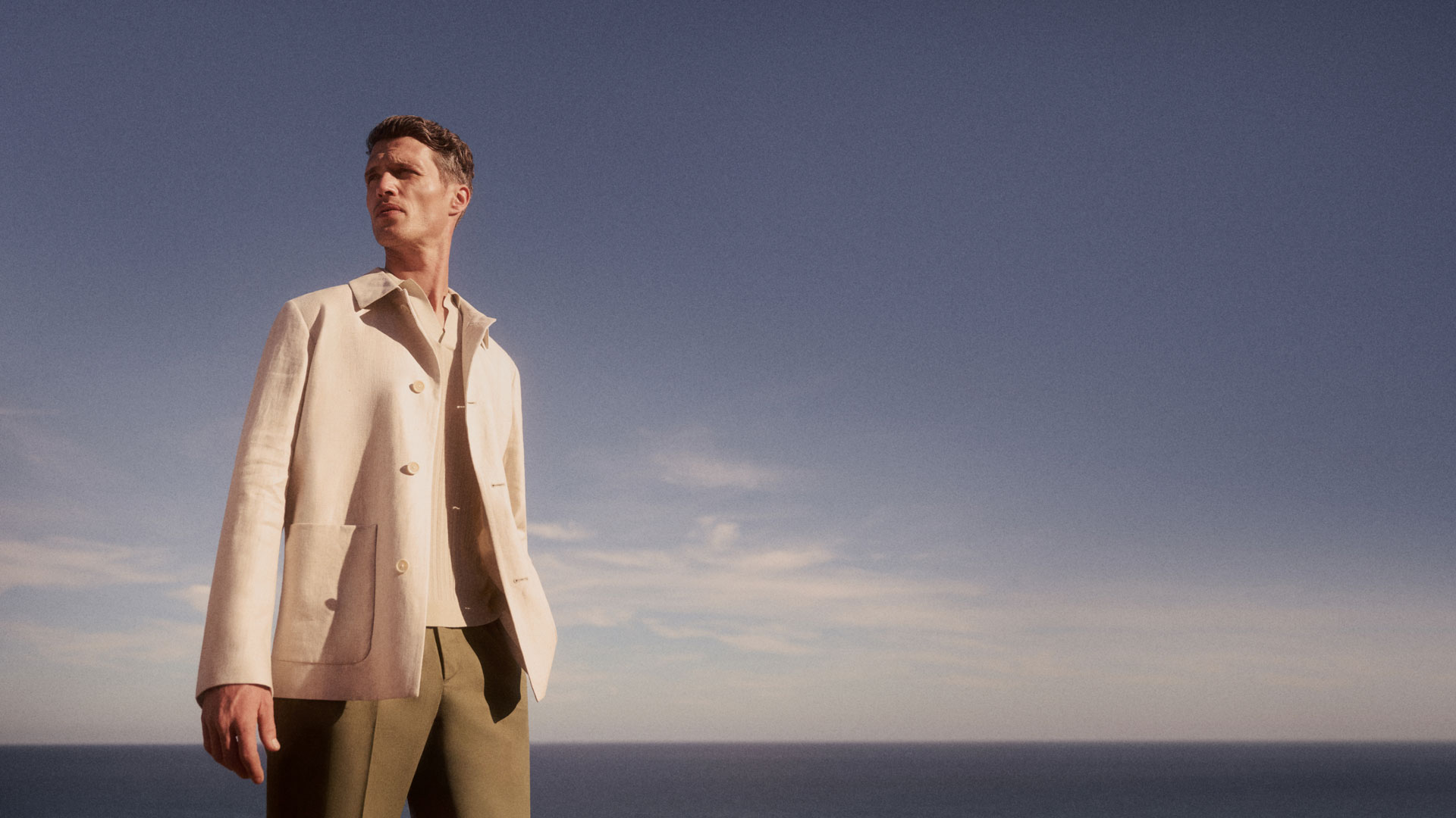 Man wearing a beige jacket and green pants standing outdoors with a clear blue sky and ocean in the background. Man wearing a beige jacket and green pants standing outdoors with a clear blue sky and ocean in the background.