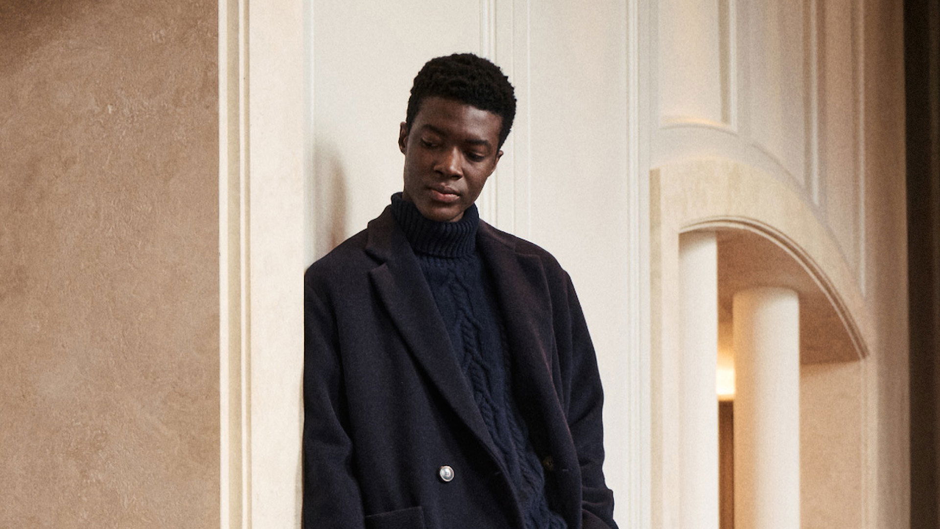 Junger Mann mit dunklem Mantel und blauem Rollkragenpullover lehnt an heller Wand in elegantem Innenraum. Junger Mann mit dunklem Mantel und blauem Rollkragenpullover lehnt an heller Wand in elegantem Innenraum.